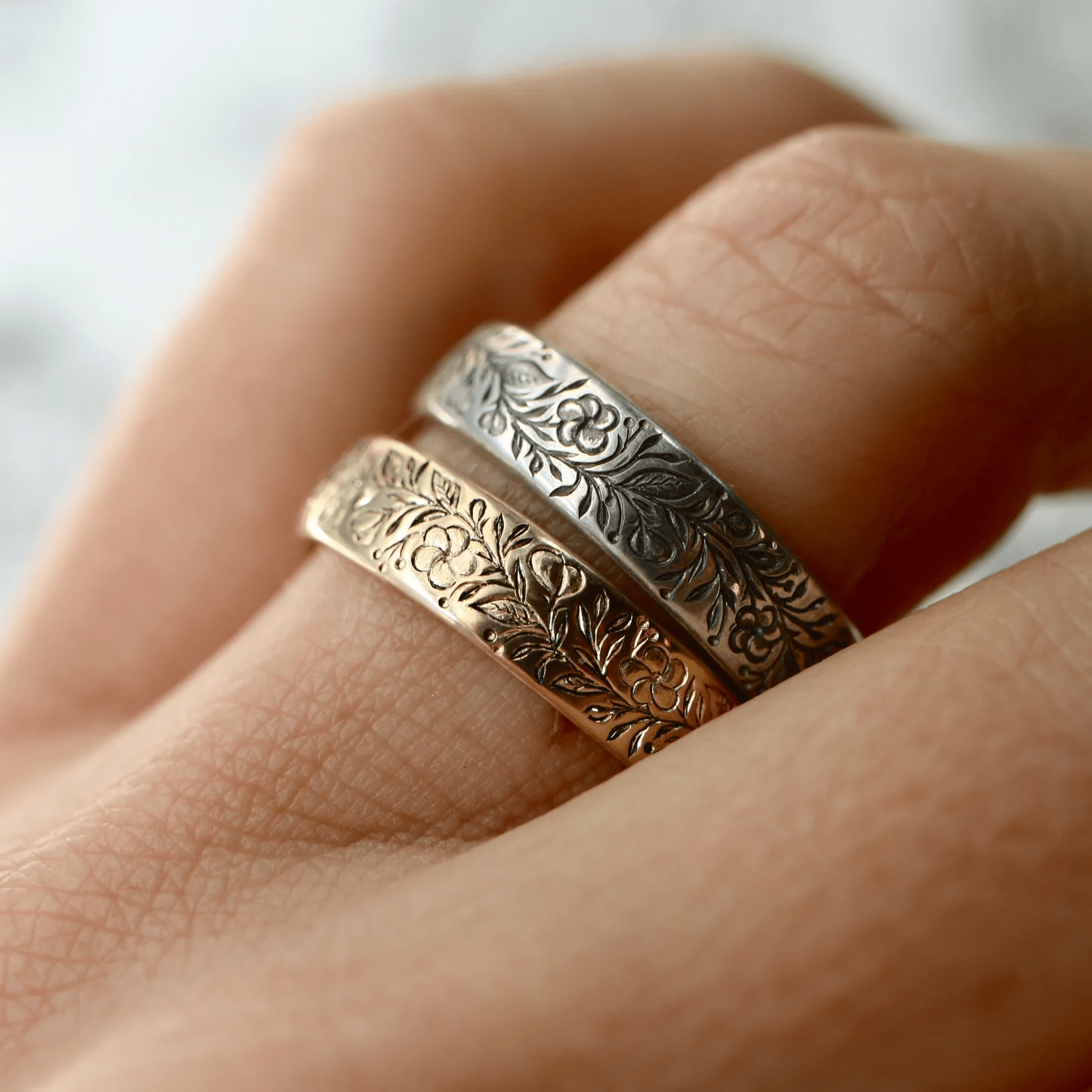 Mixed-metal engraved floral band rings worn on a hand, showing detailed floral engraving.