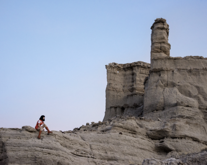 Space Waitress by Nicole Cudzilo, an archival pigment photography print featuring a small figure in red beside a towering sculptural desert rock formation.
