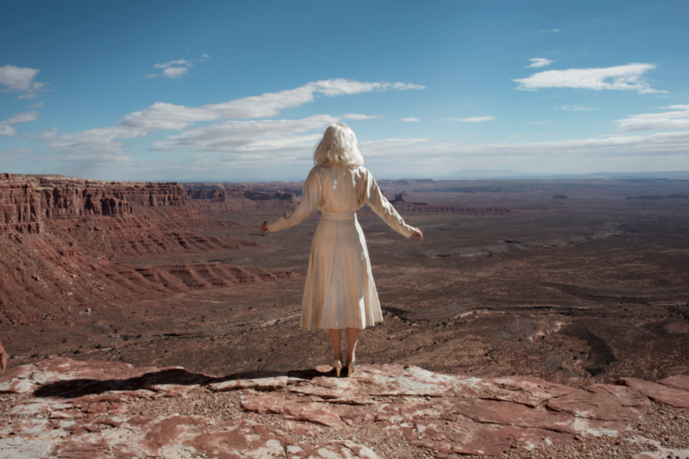 The Edge by Nicole Cudzilo, an archival pigment photography print showing a solitary figure in a pale dress overlooking a vast red rock desert landscape.