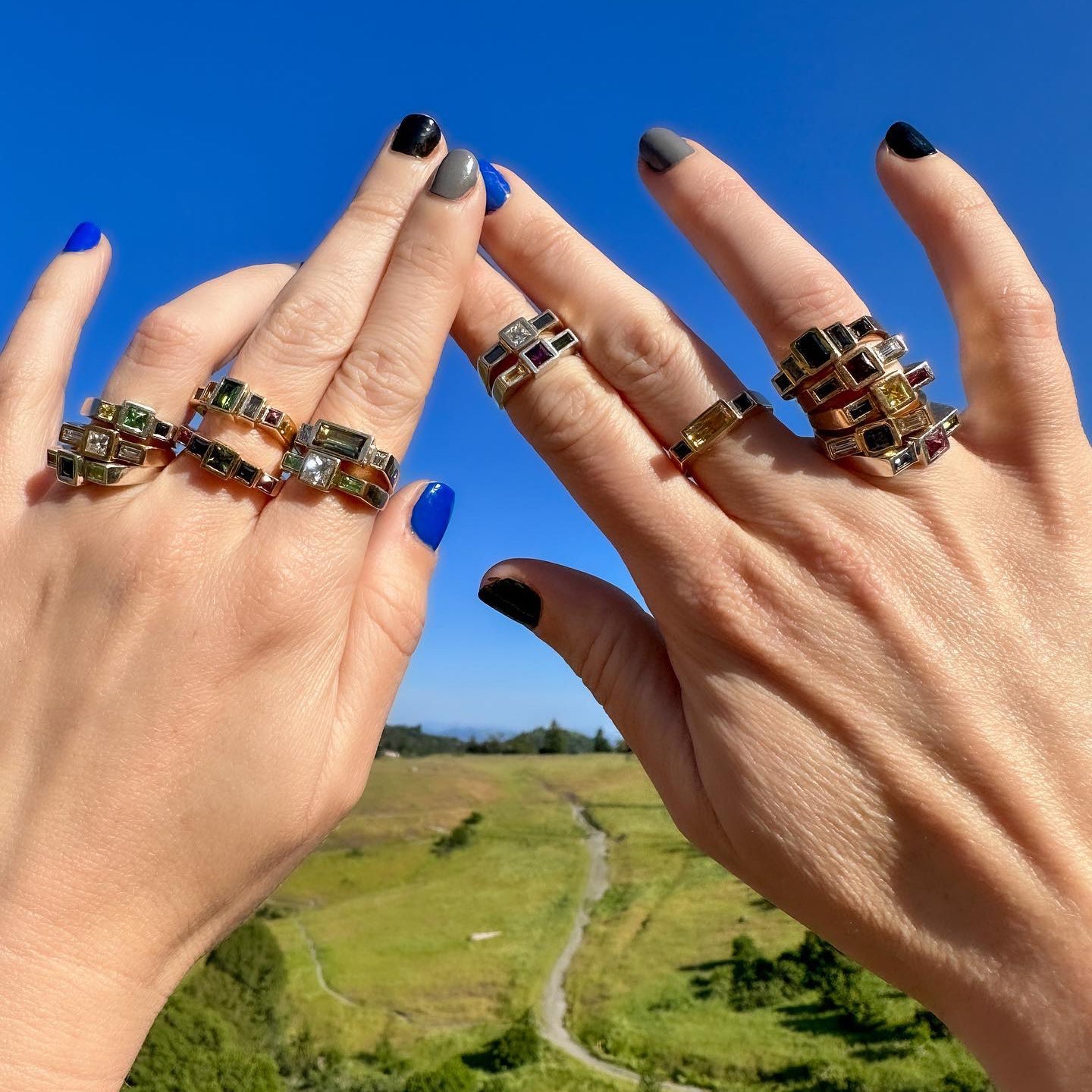 Two hands wearing multiple gemstone rings outdoors with a bright blue sky and green landscape in the background