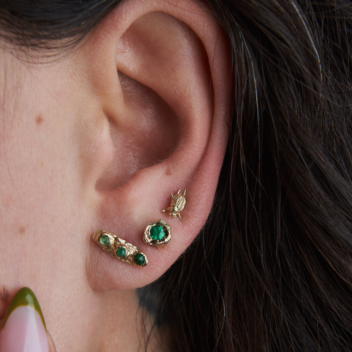 Close-up of Emerald Petra Stud Earrings worn on the ear, showing their petite 5mm size, vivid green emeralds, and textured 14k yellow gold settings.