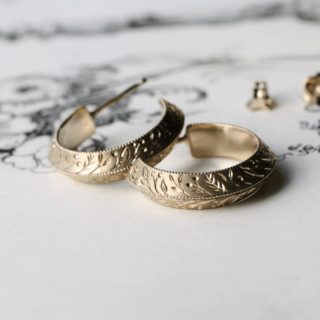 Close-up of 14k gold hand-engraved floral hoop earrings on a white background.