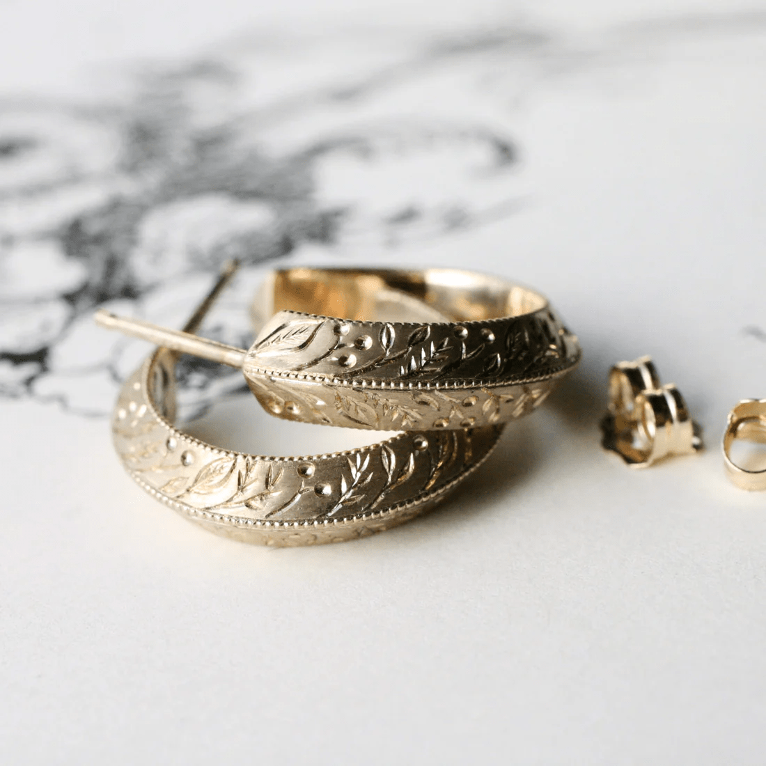 Front-facing view of 14k gold floral engraved hoop earrings on a white illustrated surface.