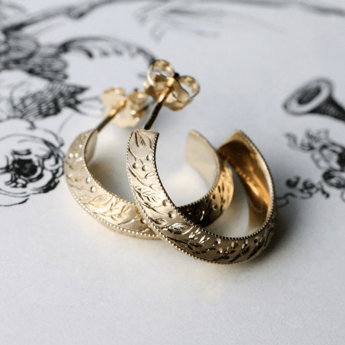 Pair of 14k gold floral engraved hoop earrings arranged together on a white background.