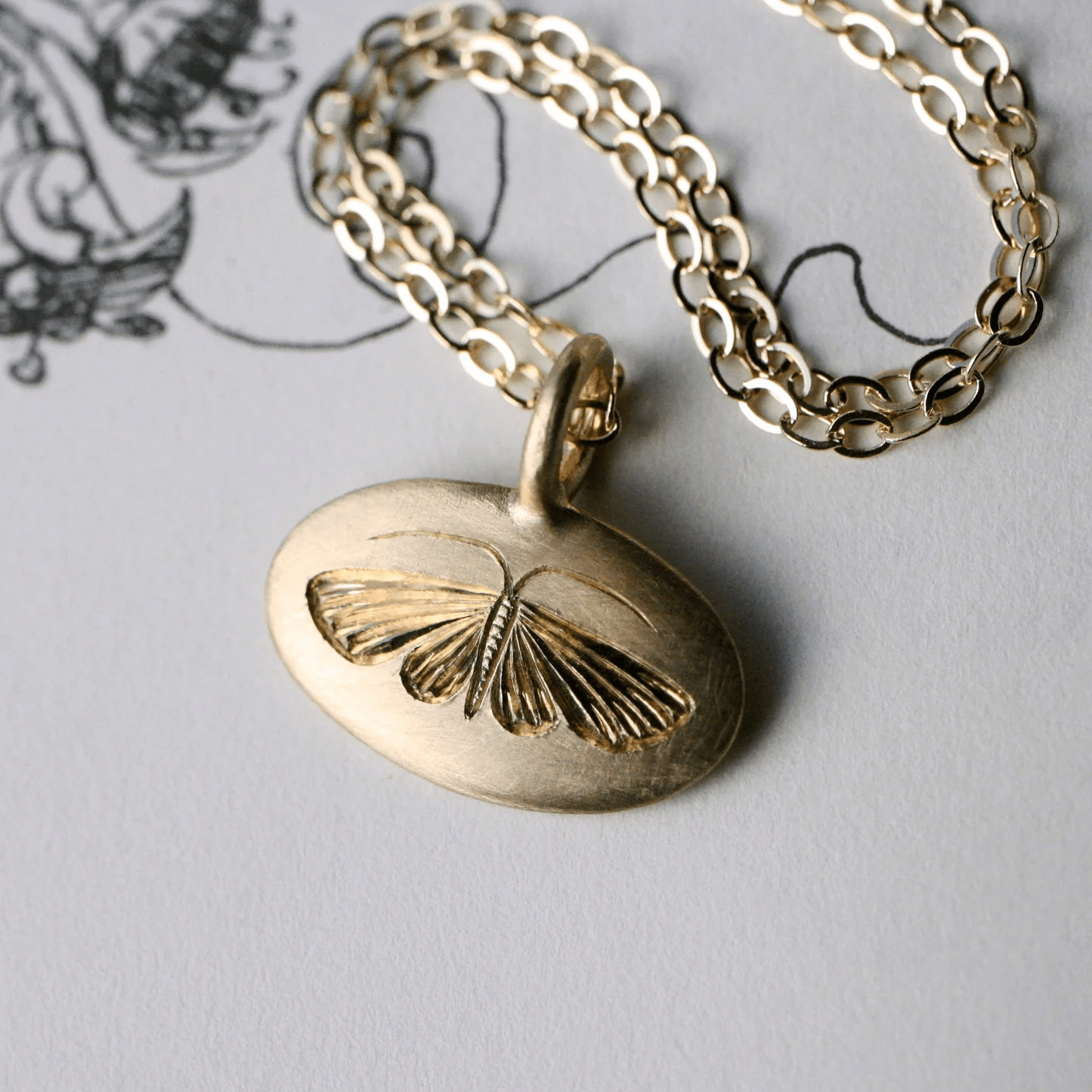 Close-up of a 14k gold hand-engraved house moth pendant on a chain.