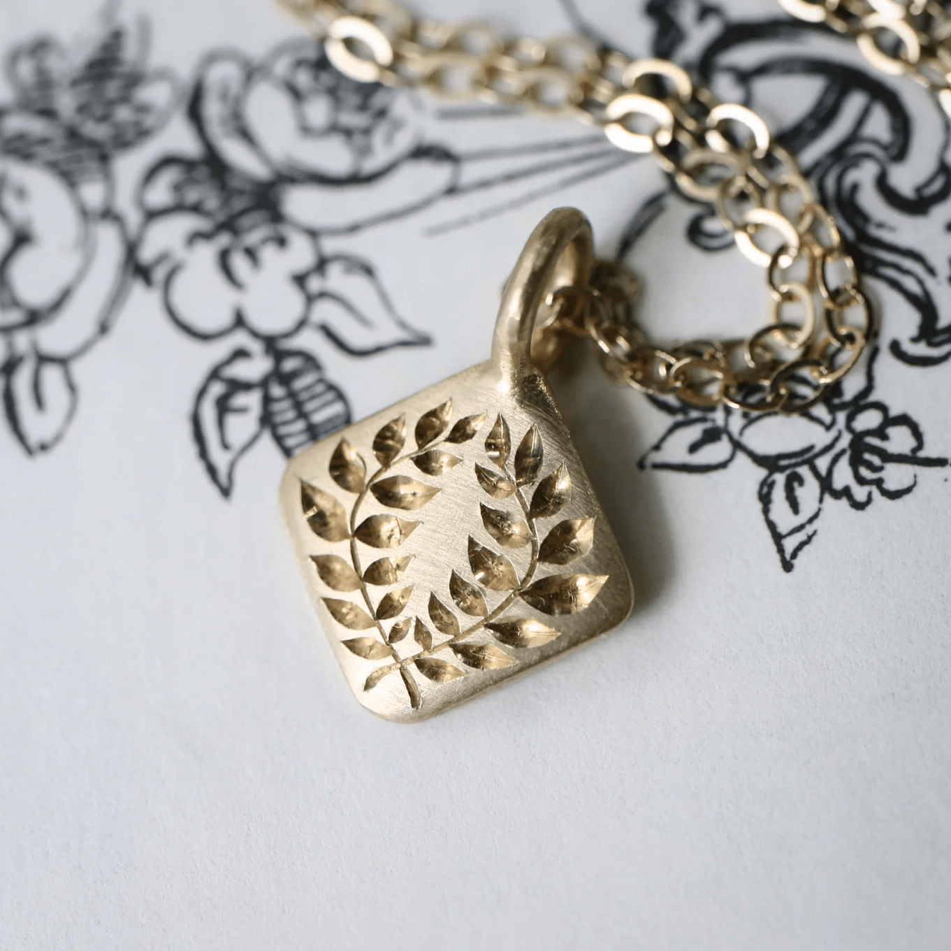 14k gold laurel square pendant with hand engraved details on a chain, shown in close-up on a white illustrated background.