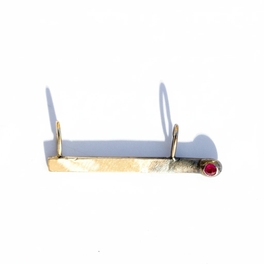 Gold match charm with a red ruby on a white background