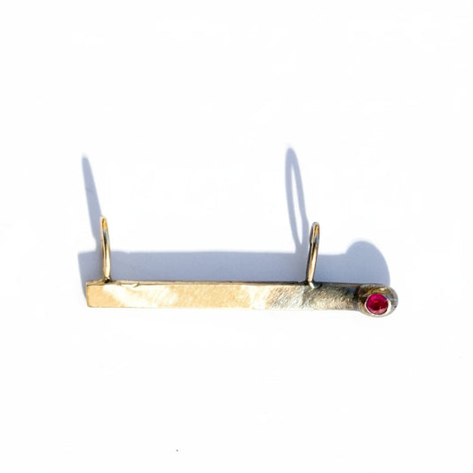 Gold match charm with a red ruby on a white background