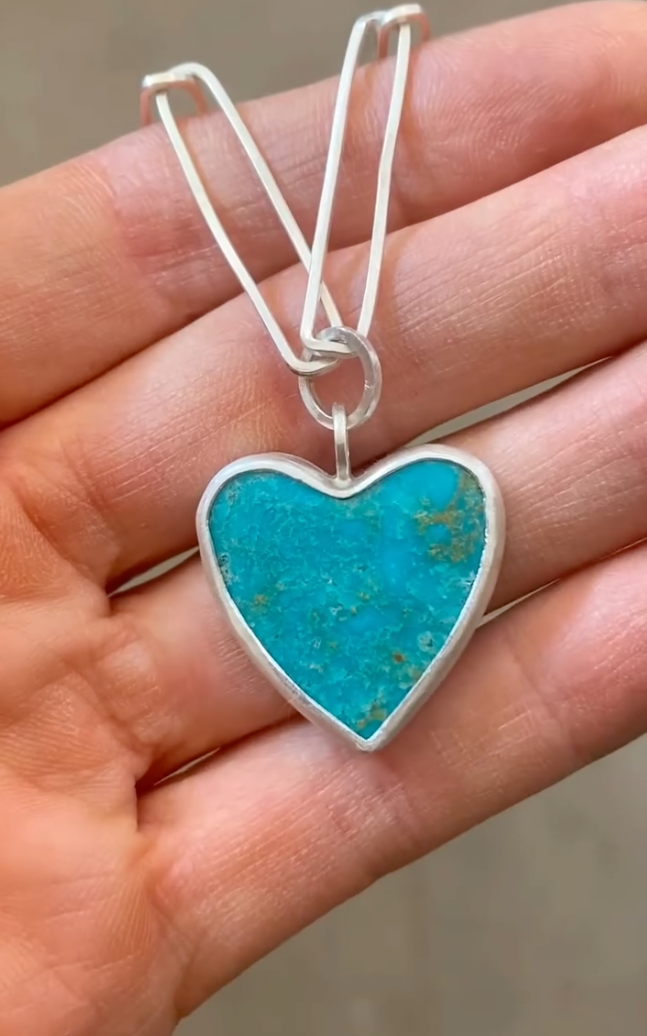 Turquoise heart-shaped pendant on a silver chain held in a hand.