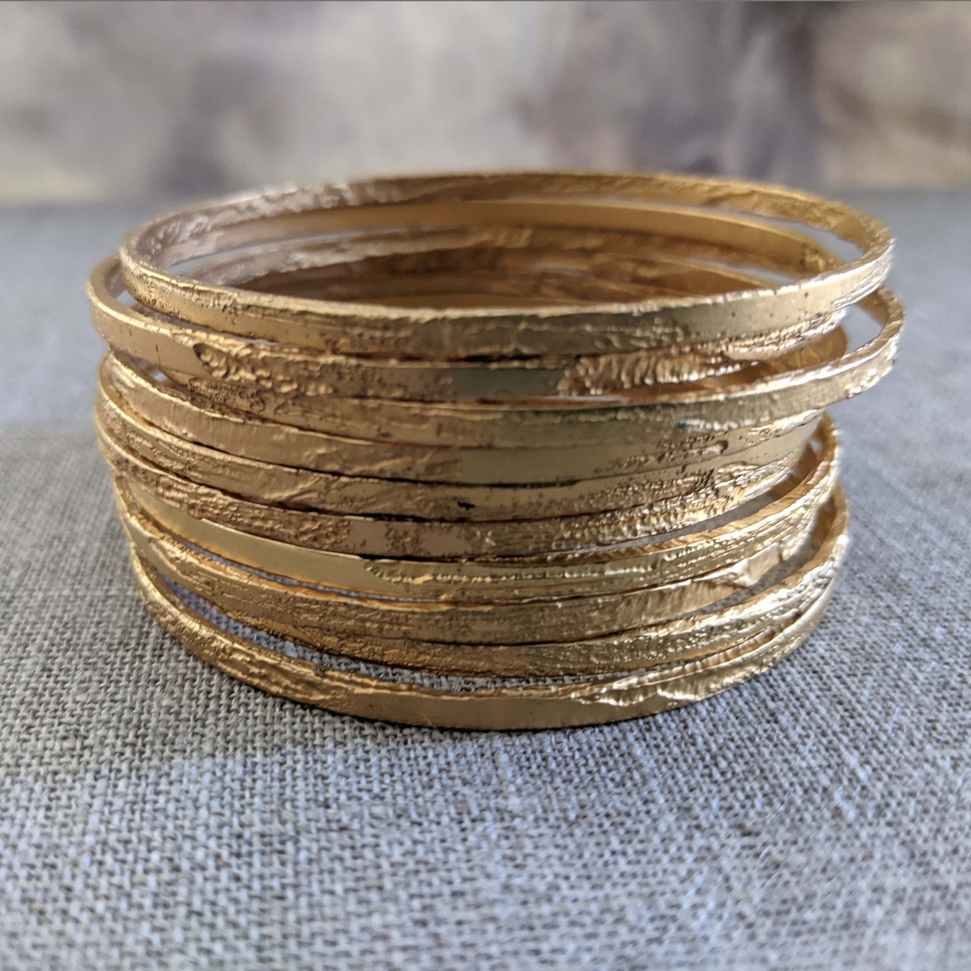 Thin etched gold bangles stacked together on a neutral textured surface.