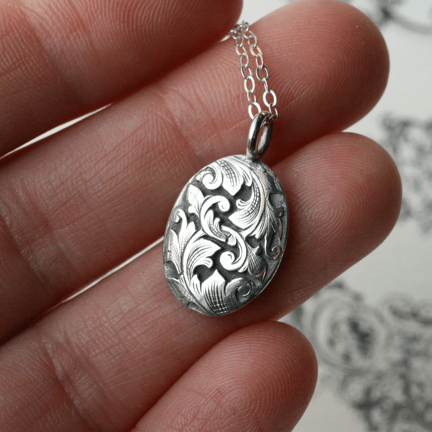 Hand-engraved sterling silver Acanthus oval pendant by Blue Blue Driver shown held between fingers.