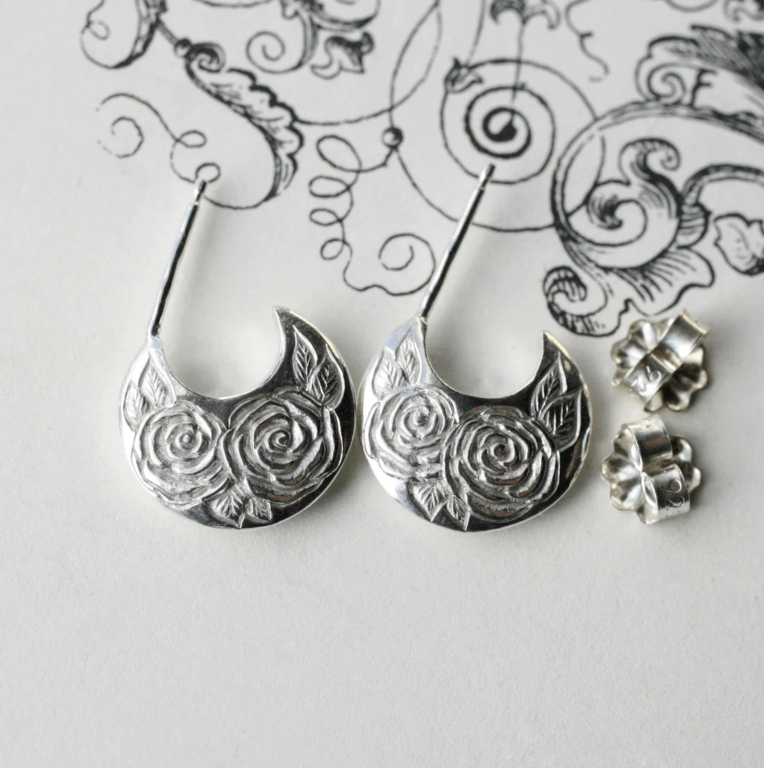 Sterling silver Beloved Rose disc hoop earrings by Blue Blue Driver displayed on a decorative sketch background with matching earring backs.