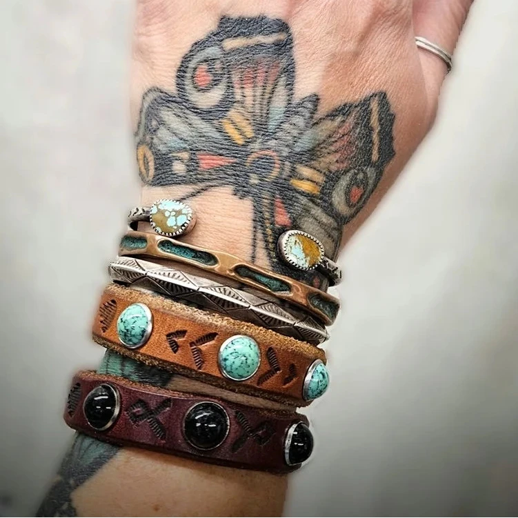 Turquoise-studded brown leather cuff bracelet styled with other handmade bracelets on a tattooed wrist.