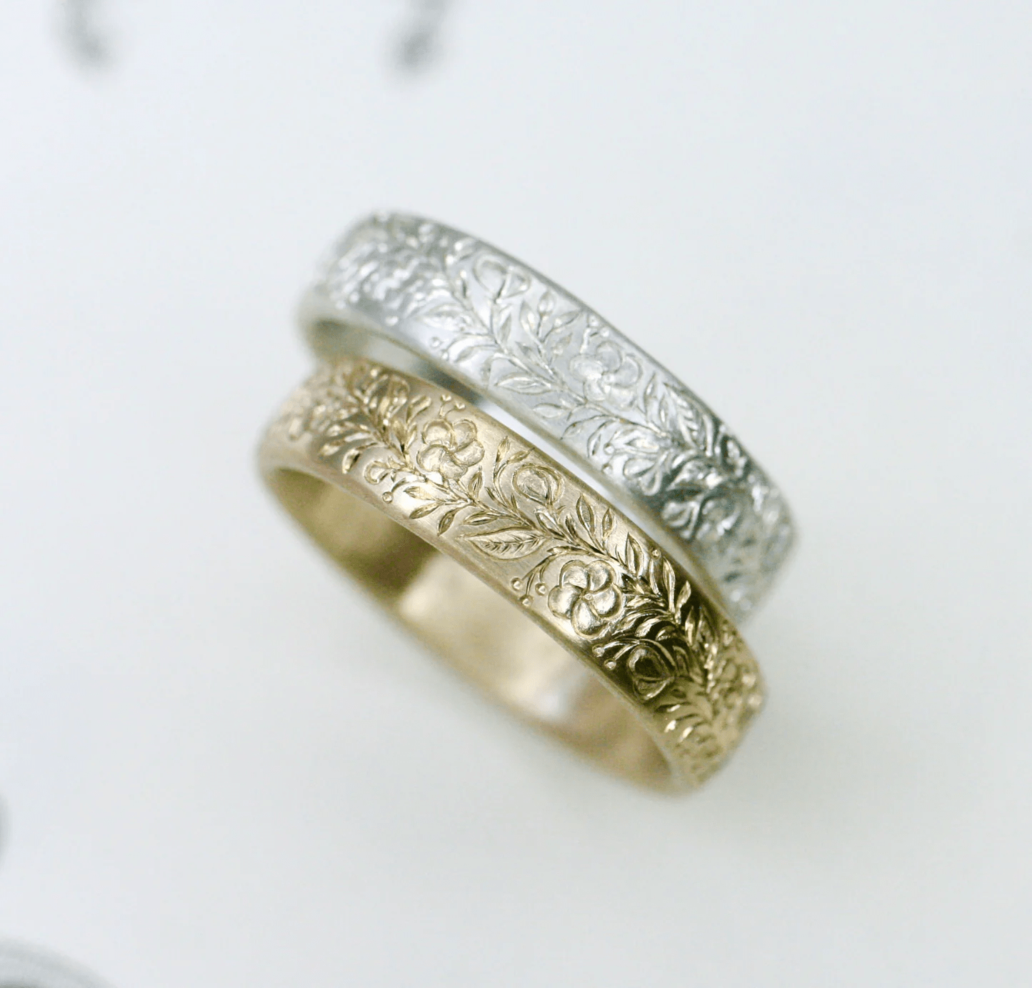 Stacked gold and white hand-engraved floral band rings displayed on a light background.