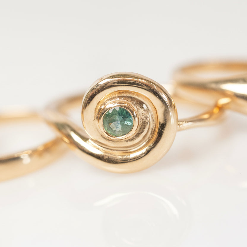 Close-up of the teal sapphire gemstone surrounded by a swirling 14k yellow gold spiral setting, highlighting the polished metal and sculptural details.
