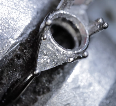 Macro photograph of a metal ring setting held in a jeweler's clamp, showing the open gemstone seat and surrounding metalwork.