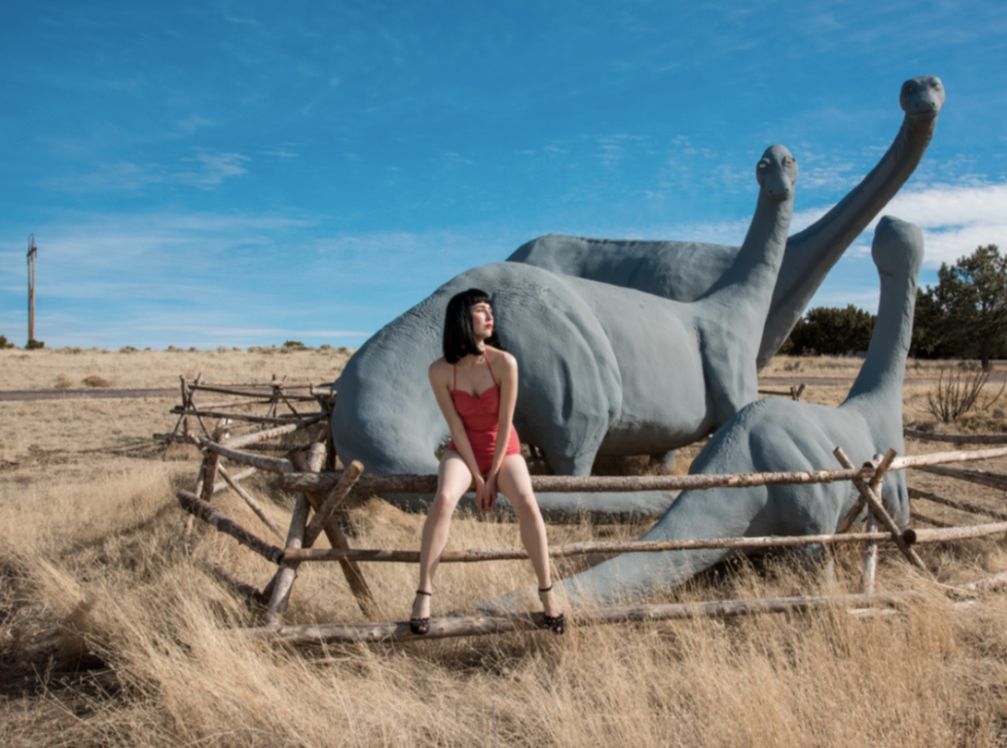 Dino Fence by Nicole Cudzilo, an archival pigment photography print featuring a figure in red beside a blue-gray dinosaur sculpture behind a rustic desert fence.
