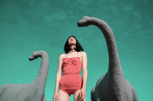 Dinos by Nicole Cudzilo, an archival pigment photography print featuring a woman in pink among towering dinosaur sculptures against a teal backdrop.