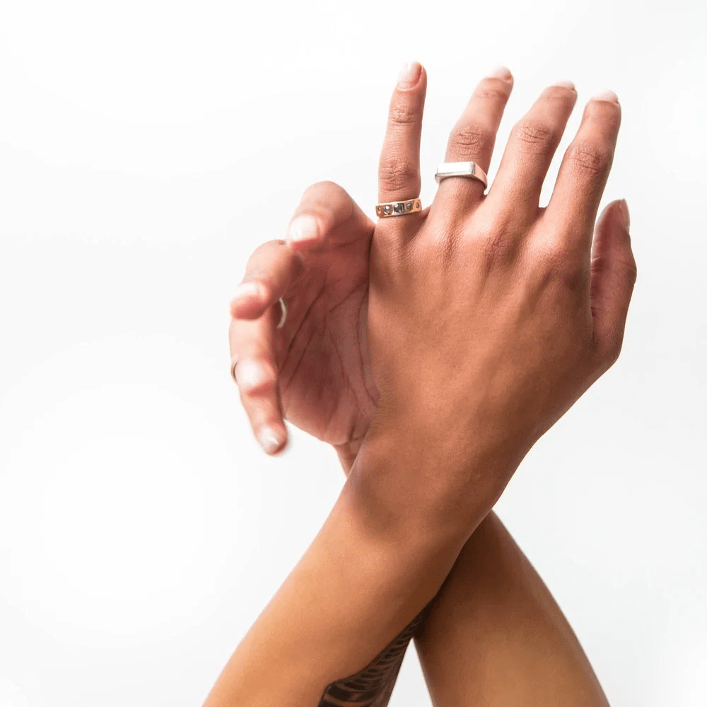 Two hands wearing multiple gold rings, featuring the Quintet Ring with five hexagon diamonds.