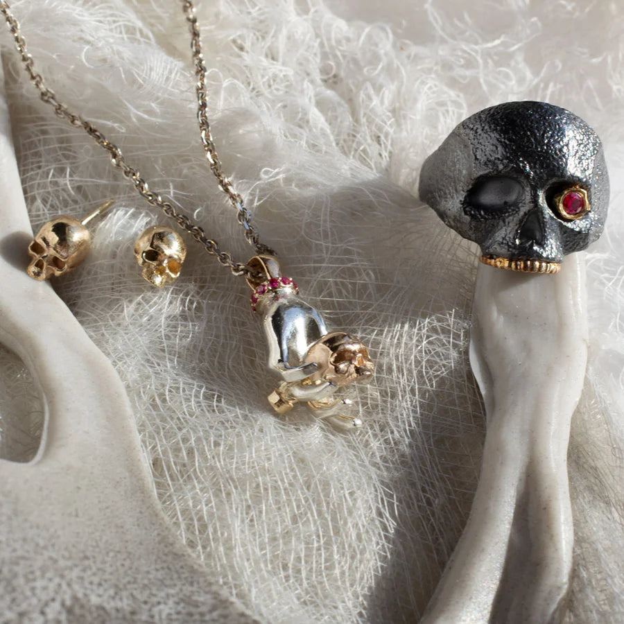The Masque of the Red Death Skull Ring displayed on a fabric surface, showing its oxidized silver finish, ruby eye, and 14k gold accents.