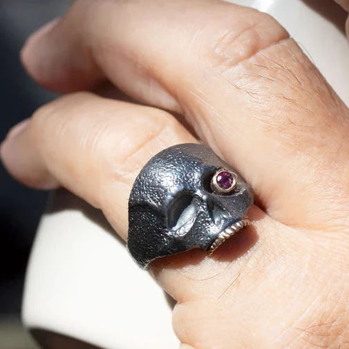 Close-up of The Masque of the Red Death Skull Ring worn on a finger, highlighting its dramatic silver skull, ruby eye, and gold accents.