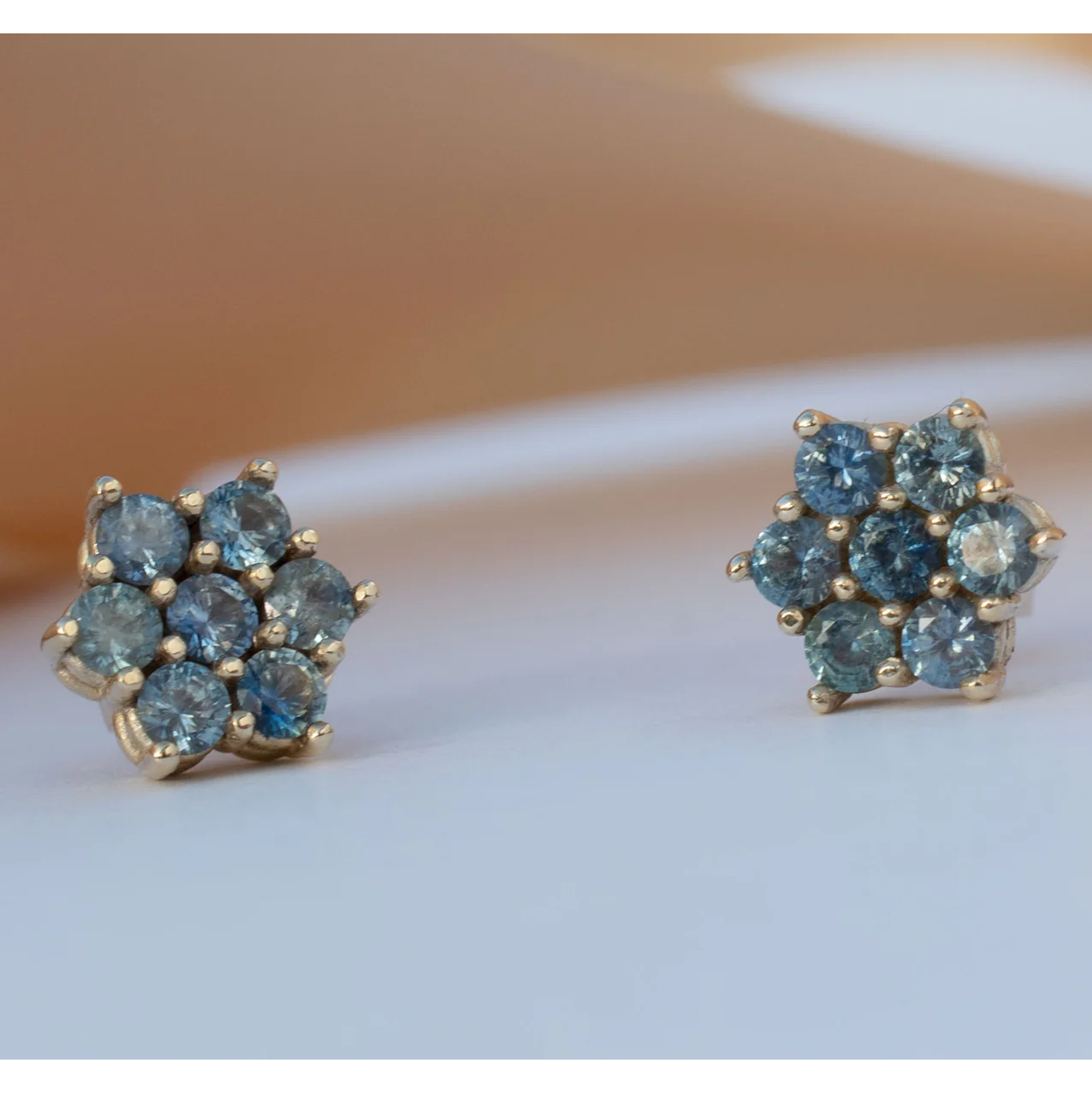 14k gold rose-shaped stud earrings with blue Montana sapphires photographed on a ceramic surface.