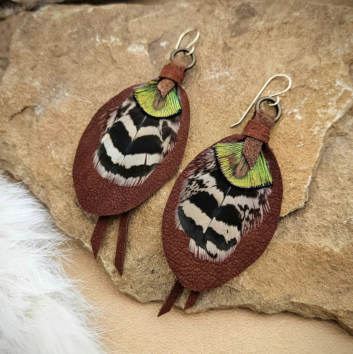 Russet leather and feather earrings displayed on natural stone, featuring layered black-and-cream feathers, iridescent green details, and gold-filled ear wires.