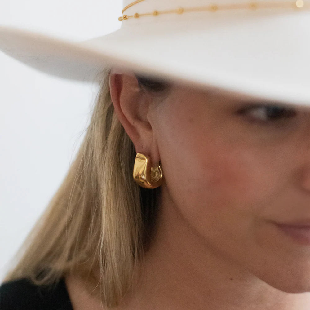 Profile view of model in a hat wearing Serrano Hoops in 14k gold, minimal sculptural hoops.