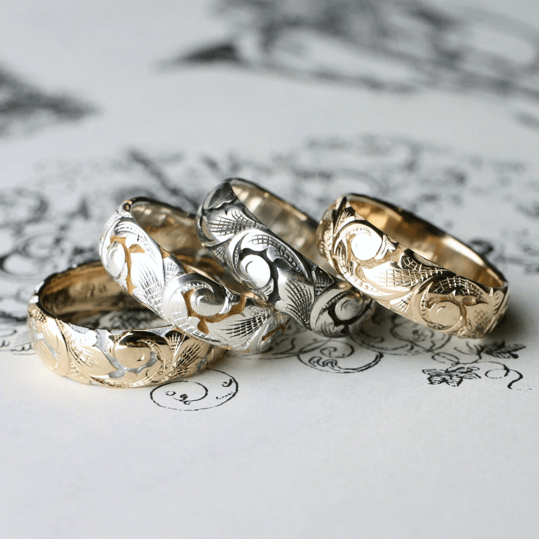 Group of slim acanthus engraved bands in gold and two-tone silver on decorative background.