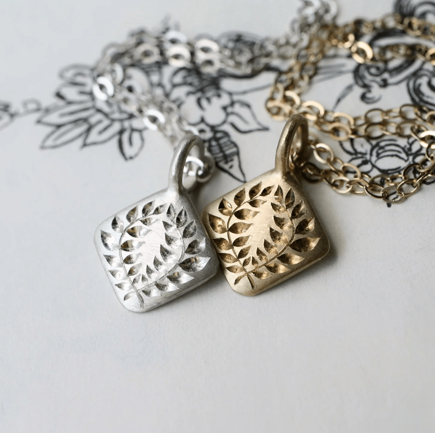 Sterling silver and 14k gold laurel square pendants displayed together on illustrated paper with matching chains.