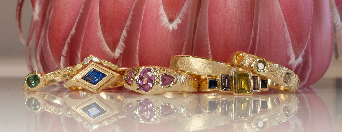 Handcrafted gold rings with colorful gemstones arranged on a reflective surface in front of a pink floral backdrop at Teko Gallery.