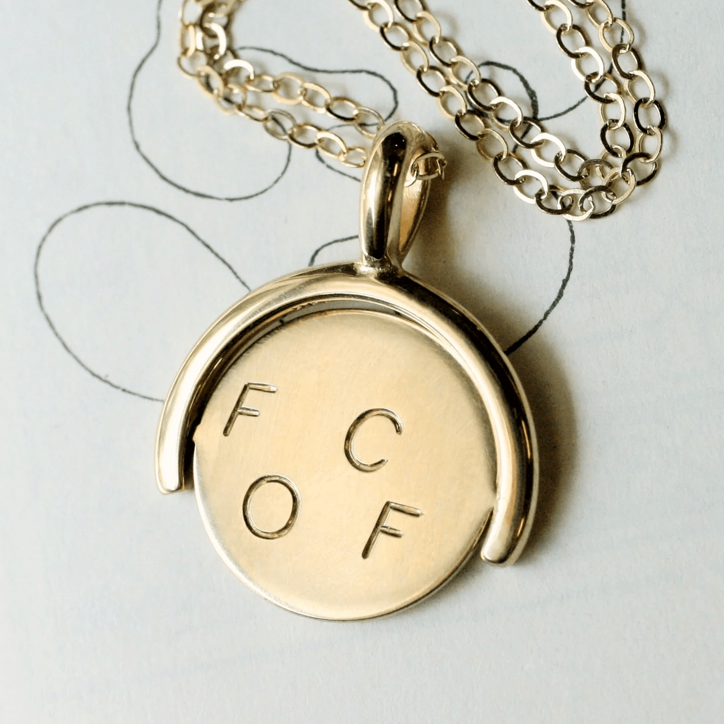 14k gold thaumatrope pendant shown from the side, featuring the engraved letters F, C, O, F on each half-disc, displayed on a sketchbook background with a gold cable chain