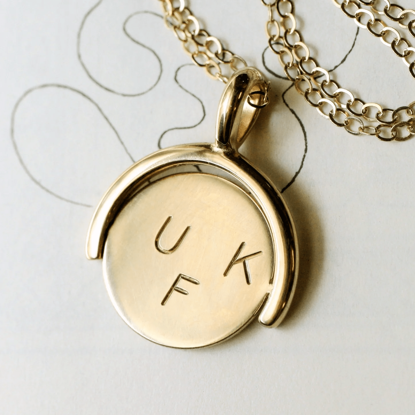 Close-up side view of the 14k gold thaumatrope pendant engraved with U, K, F, placed on a sketchbook background with a gold cable chain.