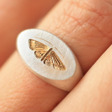 Two-tone house moth ring with a sterling silver band and gold engraved moth, worn on a finger.